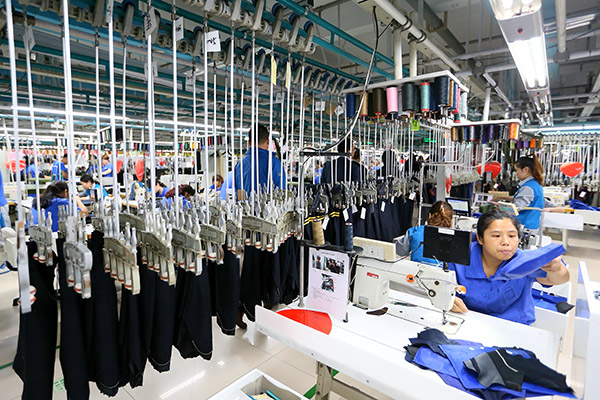Clothing factory with workers at sewing machines, garments on overhead racks.